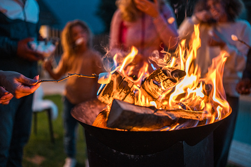 A diverse family engages in toasting marshmallows over a fire pit outside their home, enjoying a leisurely bonding activity together in casual attire.