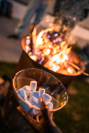 A close-up of a person's hand holding a bowl filled with fluffy marshmallows, ready to be toasted over a blazing bonfire during a casual outdoor gathering.