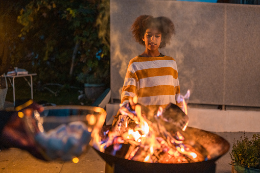 A focused young woman of diverse ethnicity delights in toasting marshmallows over glowing flames at an outdoor fire pit, her casual striped attire suggesting a relaxed evening.