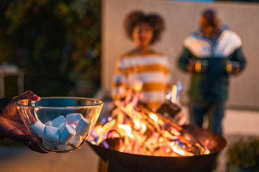 A diverse family enjoys a casual outdoor gathering. In the foreground, a hand holds a bowl of marshmallows ready for toasting over a warm fire pit, as adults chat joyfully in the background.