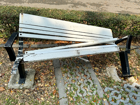 Stock photo showing a close-up view of vandalised public park bench with twisted metal leg and armrest, and lifted wood seat slats.