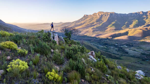 Female trail runner silhouette standing on mountain peak looking out at mountain range at sunset