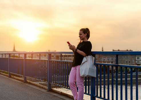 Urban carefree woman stands on the bridge while sunset and messaging online using her mobile phone. Urban lifestyle.