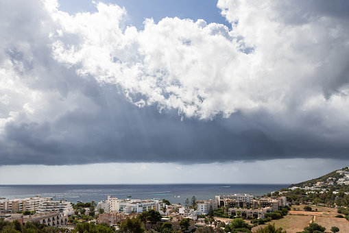 A coastal city in Ibiza under a dramatic sky, with dark storm clouds gathering above, contrasting sharply against the bright Mediterranean backdrop, capturing a moment of natural suspense