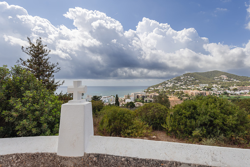 View from Puig de Missa church, featuring a symbolic white cross with the panoramic backdrop of Santa Eulalia town, its verdant landscapes, and the sparkling Mediterranean, framed by a dynamic cloud-filled sky