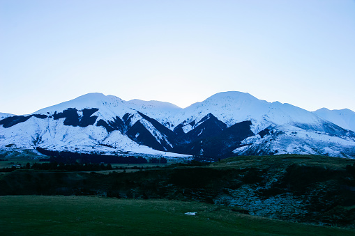 Mountains covered in snow sit on top of green pastures at sunset with a clear blue sky