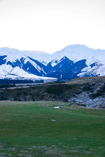 Mountains covered in snow sit on top of green pastures at sunset with a clear blue sky