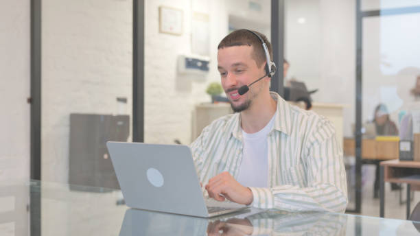 istock Creative Young Man with Headset Doing Video Chat on Laptop in Call Center 2148496008