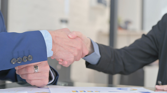 Two happy business partners in elegant suits shaking hands against workplace in office