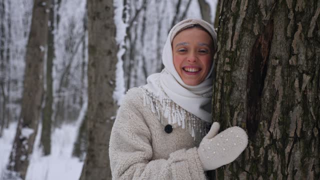Woman in white scarf and coat standing in snowy forest. Lady standing in snow-covered woods. Winter scene with woman in white coat walking in the winter forest. Female in snowy forest landscape. Snowy forest backdrop with woman in white coat.