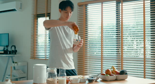 Young Asian man drizzling honey over a bowl of muesli and fruit at modern home. Diet and healthy food morning routine.
