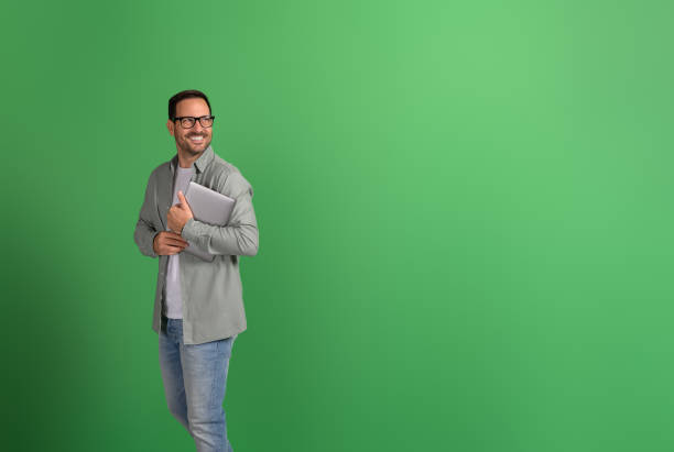 istock Positive businessman holding laptop and looking away thoughtfully while standing on green background 2148497835