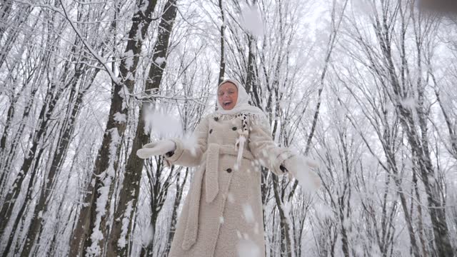 Woman in white scarf and coat throws snow high up in the forest. Lady standing in snow-covered woods. Winter scene with woman in white coat walking in the winter forest. Female in snowy forest landscape. Snowy forest backdrop with woman in white coat.