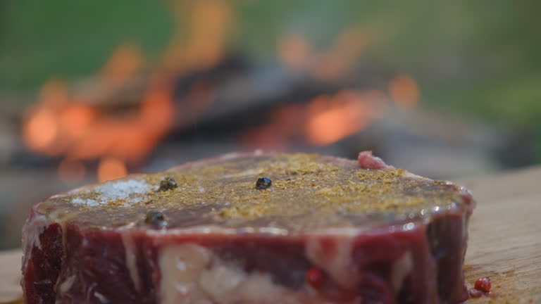 Close up of male hand sprinkling salt and spices on raw steak on wooden board near bonfire. Cooking food over campfire. Camping vibes and outdoor lifestyle mood concept
