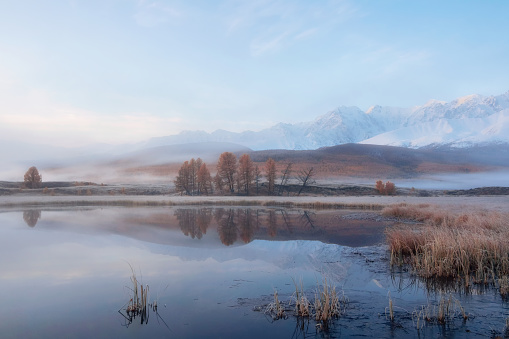 Blue hour in the mountains. Autumn frost on dry grass. Snowy peaks are reflected in the calm cold water of a mountain lake. Natural snowy mountain minimalist landscape. Perfect image for wall, screen
