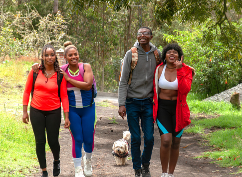 A group of four interracial friends immerse themselves very happily and cheerfully along a quiet forest path. Radiating positive energy and unity, their faces light up with laughter and smiles. A small dog accompanies them, adding to the sense of companionship and enjoyment of the outdoor adventure.