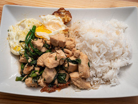 Indulge in the flavors of Thai basil chicken served with a perfectly fried egg, broccolini, and fragrant jasmine rice, elegantly presented on a white plate atop a rustic wooden board.