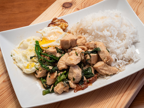 Indulge in the flavors of Thai basil chicken served with a perfectly fried egg, broccolini, and fragrant jasmine rice, elegantly presented on a white plate atop a rustic wooden board.