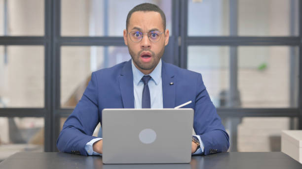 Shocked Mixed Race Businessman Looking at Camera while Working on Laptop