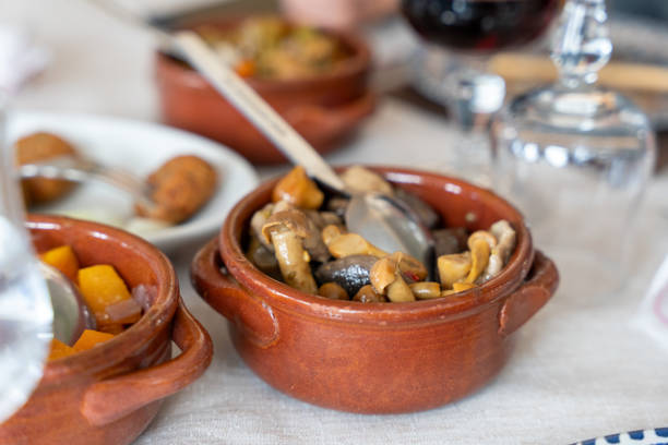 A bowl of mushrooms and other vegetables is on a table. The bowl is brown and has a spoon in it. There are other bowls on the table as well