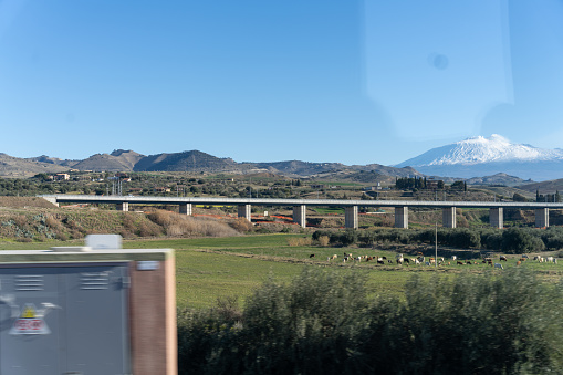 A bridge spans a valley with a mountain in the background. The sky is clear and the sun is shining
