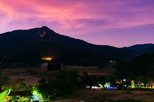 A beautiful mountain range with a purple sky in the background. The mountains are covered in trees and there are houses in the valley below. The sky is a mix of pink and purple, creating a serene