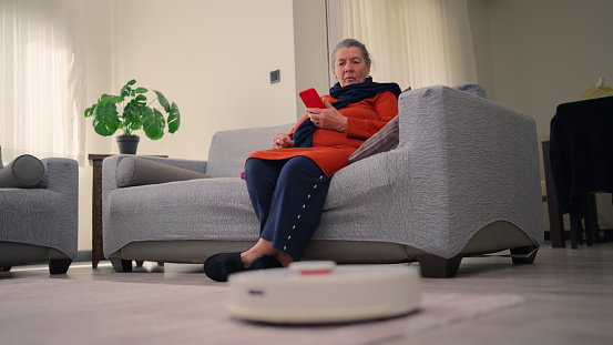 A robotic vacuum cleaner is cleaning the living room while a senior woman is sitting on a sofa and relaxing.