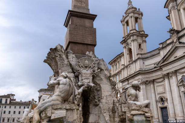 A large building with a fountain in front of it. The fountain has statues of two men and a woman. The building is old and has a lot of detail. The sky is cloudy and the sun is not visible