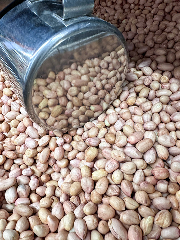 Stock photo showing close-up, elevated view of shelled peanuts pile in stainless steel container, with metal cup scoop for customers to serve themselves peanuts for healthy eating diet food snacks, nuts high in protein.