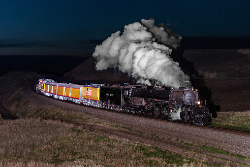 A picture of a black steam train moving on a rail track with smoke coming out of the chimney the picture is taken high above looking down