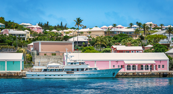 A large, white,  luxury yacht is docked in St. Georges, Bermuda with a neighborhood of colorful pastel homes in the background.