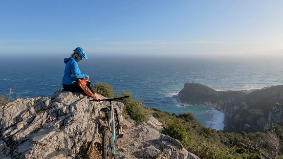 Mature woman e-mountain biker uses mobile phone on coastal viewpoint above sea, Liguria