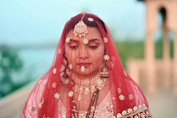 In this close-up, the exquisite features of a beautiful Indian bride are accentuated, her face adorned with a delicate veil. Despite the veil covering part of her face, her captivating eyes and serene expression shine through, radiating grace and elegance. This intimate portrait captures the beauty and charm of the bride on her wedding day