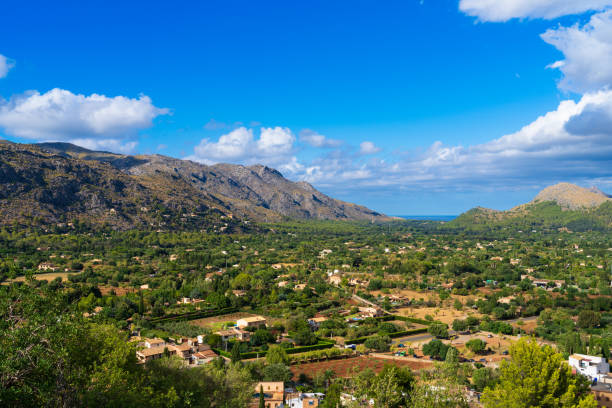 Scenic view of the Serra de Tramuntana mountain range on Mallorca, featuring rugged rock formations and rich greenery under a serene blue sky adorned with natural clouds.