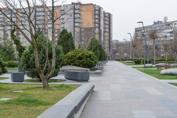 Nice modern leisure city park with benchs surrounded high-rise building. Public park in town residential. Sidewalk and walkway with conifer trees. Place to rest in city landscape. Urban architecture.