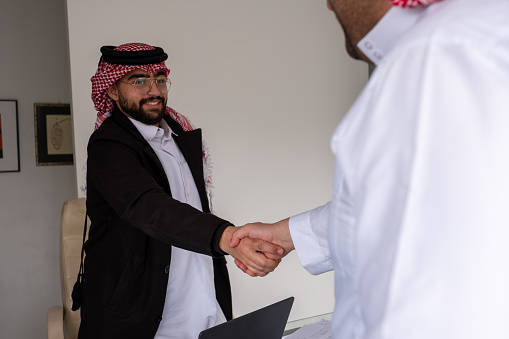 A photo of Arab businessmen making a handshake. Middle eastern professionals are making deal while sitting at conference table. Emirati man is wearing traditional clothes and other is in suit. Business partners are in Middle East office.