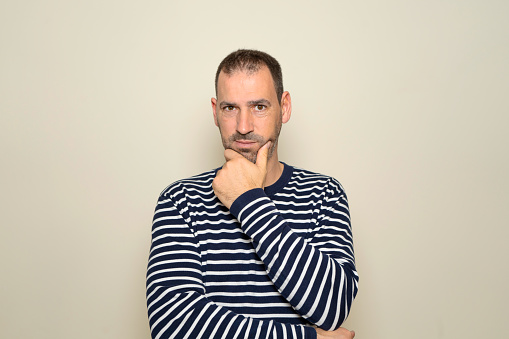 Hispanic man with beard about 40 years old wearing striped sweater thinking concentrated in doubt with finger on chin and looking at the camera wondering, isolated over beige background