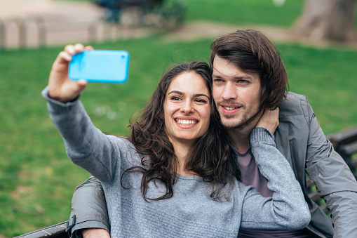 Happy young couple using smartphone outdoor in the city park. Cheerful man and woman on date sitting on the bench and taking selfie
