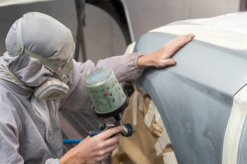 A meticulous auto painter focuses on applying an even coat of paint to a car hood in a workshop setting.