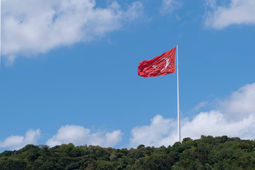 a large flag of Turkey waving on a white flagpole on a hillside with trees on a blue day, copy space, horizontal