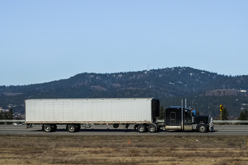 A black, classic styled semi-truck, pulling a refrigerated trailer driving on a non-urban section of the interstate freeway.