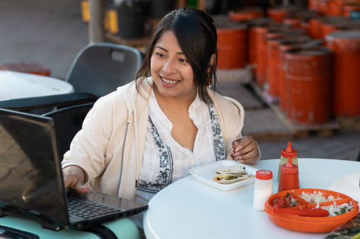 Tourist woman in her 20s, working as a freelancer with her laptop that has an internet connection, outside a taco restaurant in Mexico with a digital nomad lifestyle