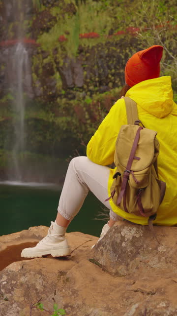 Vertical video. Woman traveler hiker in a yellow raincoat near waterfall in the highlands, enjoys nature and life. Traveling in the mountains, adventure in trip. Traveller in yellow puffer jacket