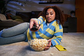 Latin woman with serious expression changing TV channel lying on room carpet with bowl popcorn.