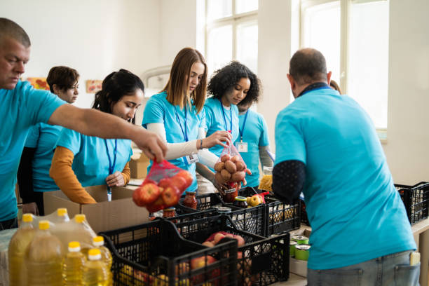 Team of charity assistants packing food contributions A young team of aid workers are busy assembling food parcels into cartons, working around a table covered with groceries and wearing matching outfits food bank stock pictures, royalty-free photos & images
