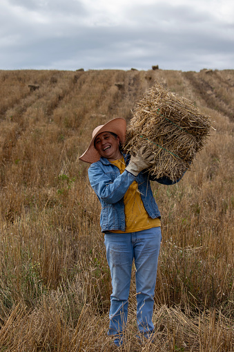 Adult woman dressed in jeans, a yellow T-shirt, and a sun hat working in a grassy field, lifting a bale of hay in the Andes Mountains