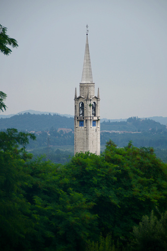 Telephoto close-up of a clock tower rising in an Italian valley, framed by trees and hills