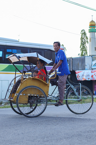 Cilacap, Indonesia - March 29, 2024 : an old man who is accompanying passengers using his pedal rickshaw service walks in the middle of the main road, while looking at the camera