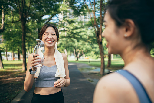 In the park, two Asian women engage in physical activities while utilizing smart devices to monitor their body data. Their interactions are filled with joy and camaraderie, creating a dynamic atmosphere amidst the natural surroundings.