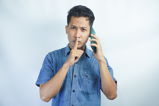 Asian man wearing blue shirt hides a secret while talking on the phone with finger on lips against white background.